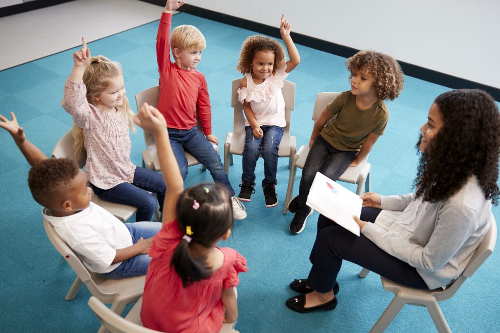 An education assistant reads aloud to her students with each of them raising their hands